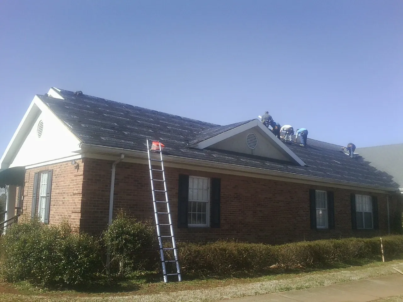 Crew tearing shingles off a brick ranch roof with a ladder set — Metal Roof Repair work by our crew in Elmira