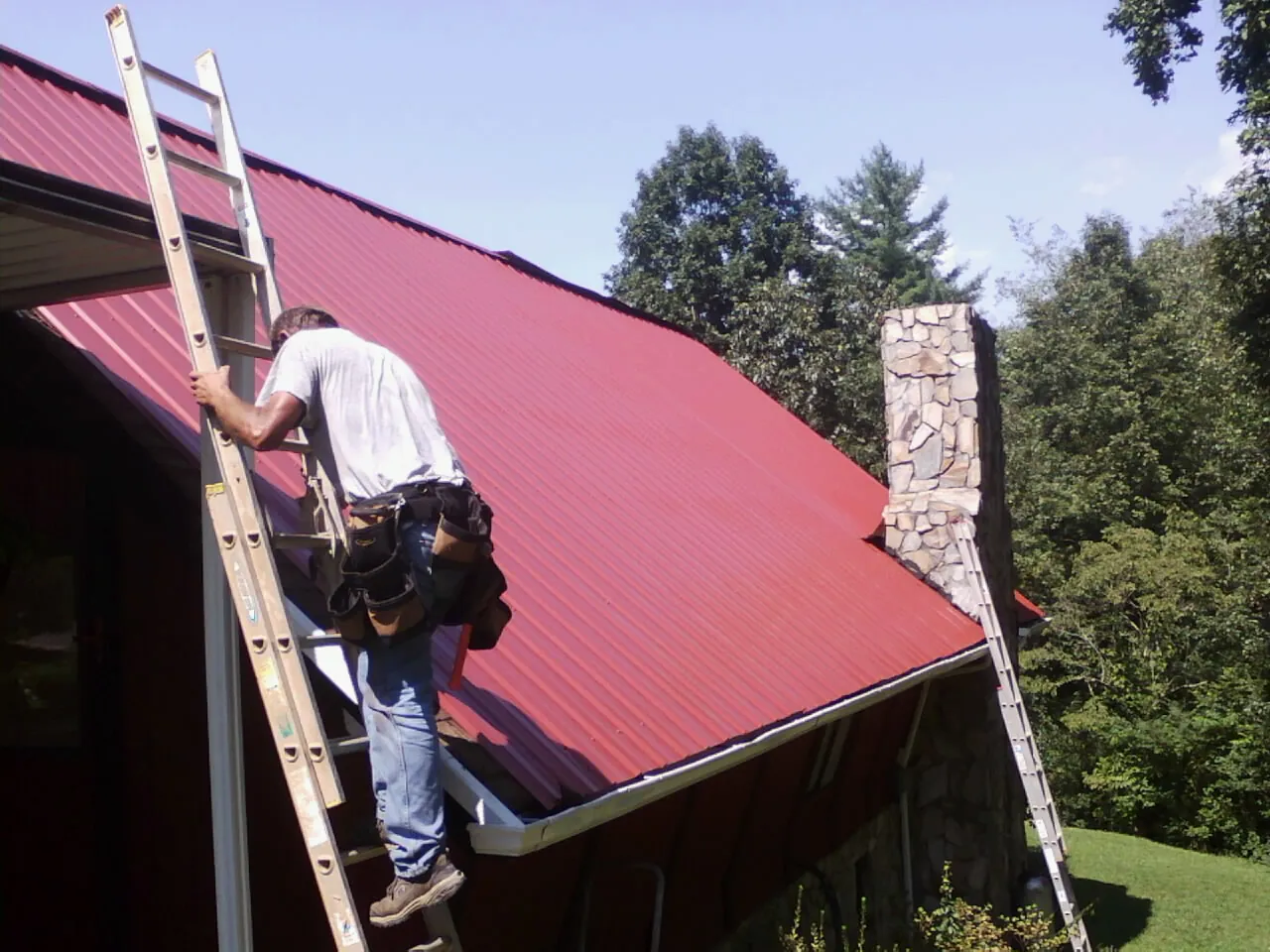 Roofer on a ladder beside a red metal roof and stone chimney — Soffit & Fascia Repair by Roofing Brothers in Elmira